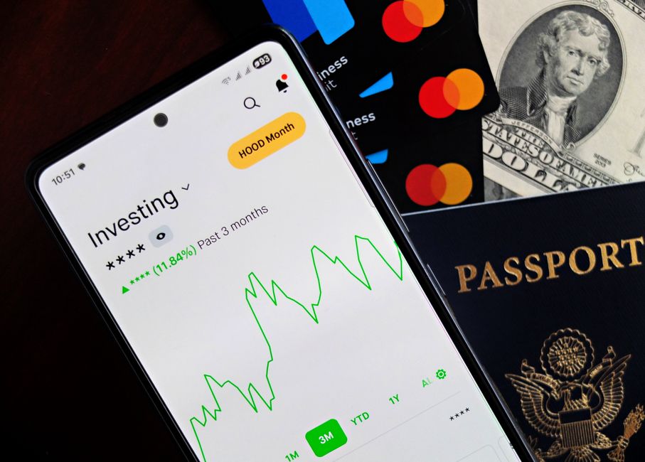 投資アプリが表示されているスマートフォン。近くにはクレジットカード、現金、パスポートがあり、金融を象徴している。