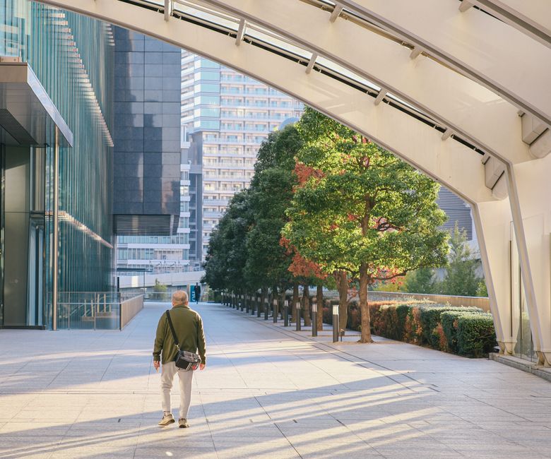 木々や高層ビルが立ち並ぶ街並みの中、一人の人が近代的なアーチの下を歩いています。