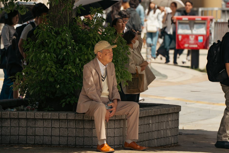 東京の街路で、歩行者に囲まれながら植物のそばに座る高齢男性。