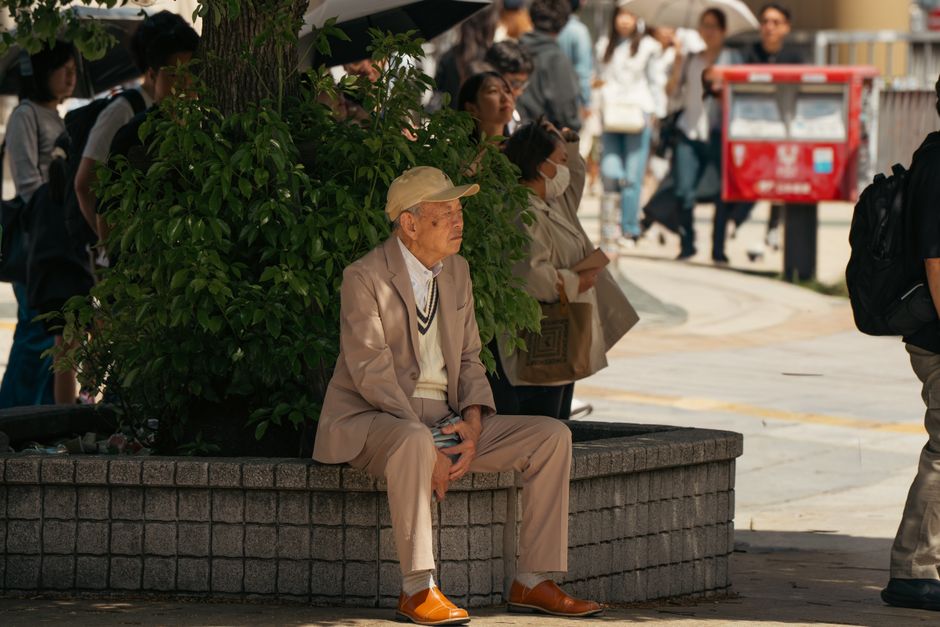東京の街路で、歩行者に囲まれながら植物のそばに座る高齢男性。