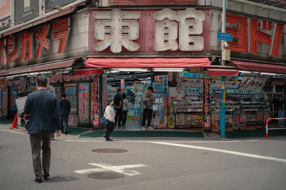 東京の賑やかな電気店の昼間の様子。店の外では人々が買い物をしている。