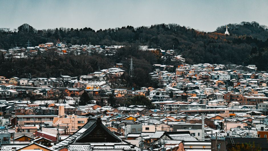 伝統的な建築と森に覆われた丘陵地帯が広がる、雪に覆われた日本の街並み。