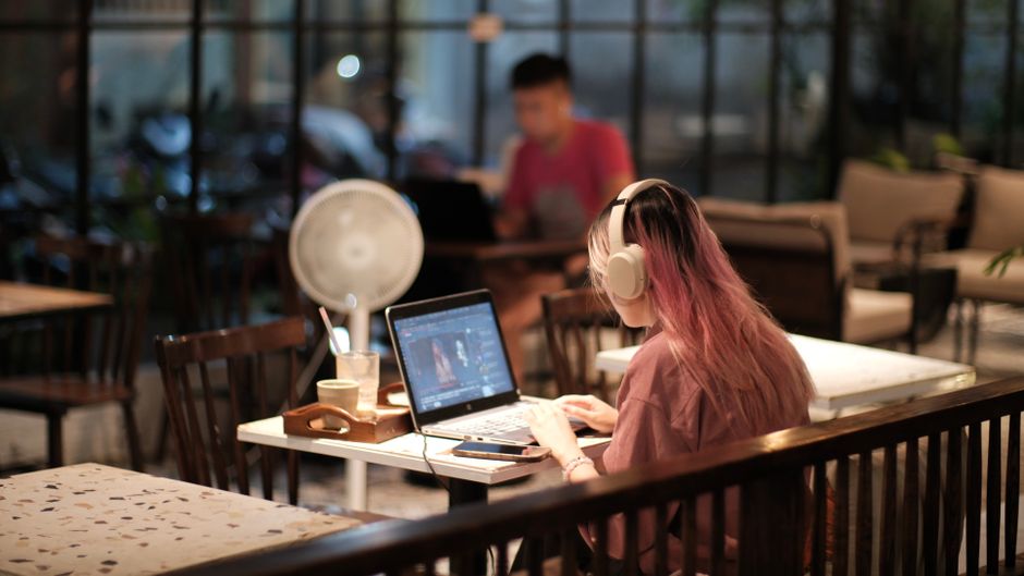 ベトナムの居心地の良いカフェで、ヘッドホンをつけた女性がノートパソコンで動画編集をしている。