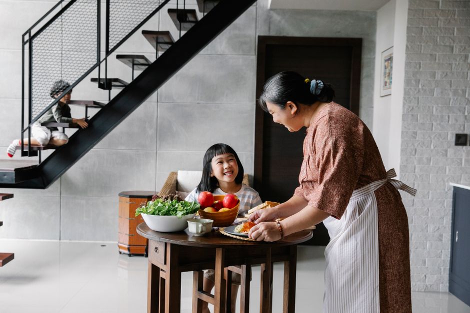 祖母が新鮮な野菜と一緒に昼食を提供している間、テーブルに座っている陽気な小さなアジアの女の子